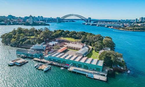 Aerial view of Goat Island in Sydney Harbour on a bright and clear day. Small boats are docked on the island, with the Harbour Bridge and Sydney skyline in the background