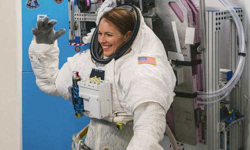 Katherine Bennell-Pegg, smiling and waving while wearing her space suit and attached to space equipment