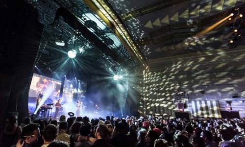 Concert-goers crowded at front of a mid-sized stage, band performing and spotlights flashing