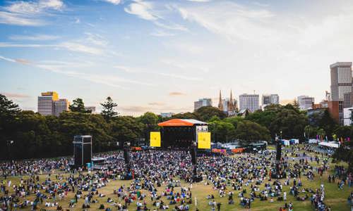 Large crowd gathered at open grass space at The Domain in Sydney, with a stage set up at the front and performances happening as the sun begins to set