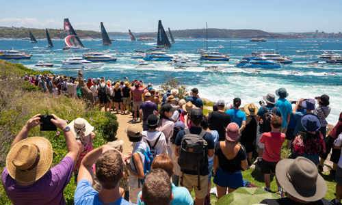 Crowd gathers by the bay in summer, taking photos and watching a fleet of yachts at the Sydney to Hobart Yacht Race