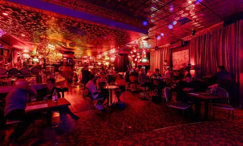 Patrons sitting at small tables in a cozy jazz club, with dim lights bouncing light on the textured ceiling