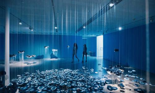 Two women walk through a dimly-lit blue room, observing an art installation of hanging strings and floating objects that are pouring to the ground