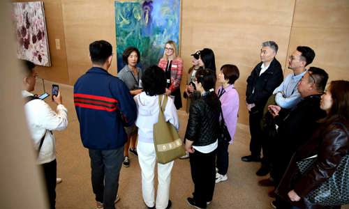 Group of 12 stopping in front of a brushed blue painting and listening to a gallery guide explain the artwork