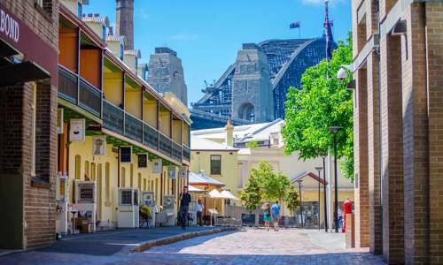 Colonial-style buildings line a cobbled lane with the Sydney Harbour Bridge visible in the distance