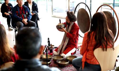 Guided sound meditation. The instructor has multiple sound bowls and is playing one while group sits in a semi circle with their eyes closed and taking in the experience